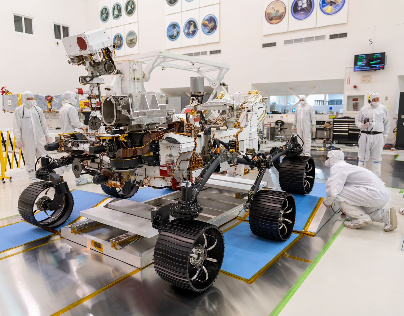 a large robotic rover with six wheels in its skeletal form being developed in a white sterile lab. the rover is about the size of a two-car door. people in white hazmat suits work on the rover