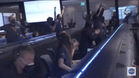 a gif recording of a group of people wearing face masks and navy blue polos in a mission control room. they all stand up and clap and cheer at the same time, celebrating the landing of perseverance rover