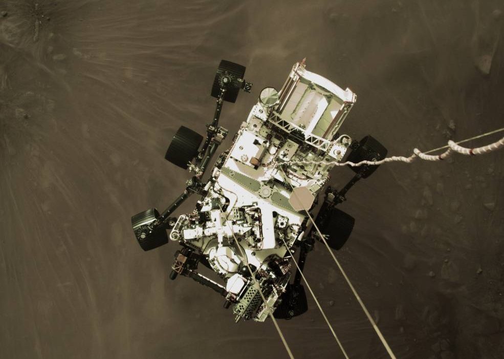 a color image aerial view of a large six-wheeled robotic rover parachuting down onto the desert environment of Mars Jezero Crater