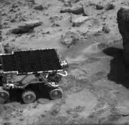 A choppy black and white movie showing a small robotic rover, named Sojourner, slowly inching towards a rock