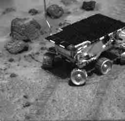 A choppy black and white movie showing a small robotic rover, named Sojourner, slowly inching towards a rock