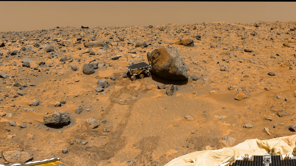 a color image of a orange dusty desert looking out at a small six-wheeled robotic rover with a solar panel on its roof. it is close to a large rock.