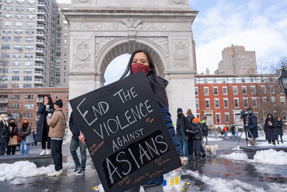 a young asian woman holds a sign that reads end the violence against asians in a snow covered plaza with an archway in the background