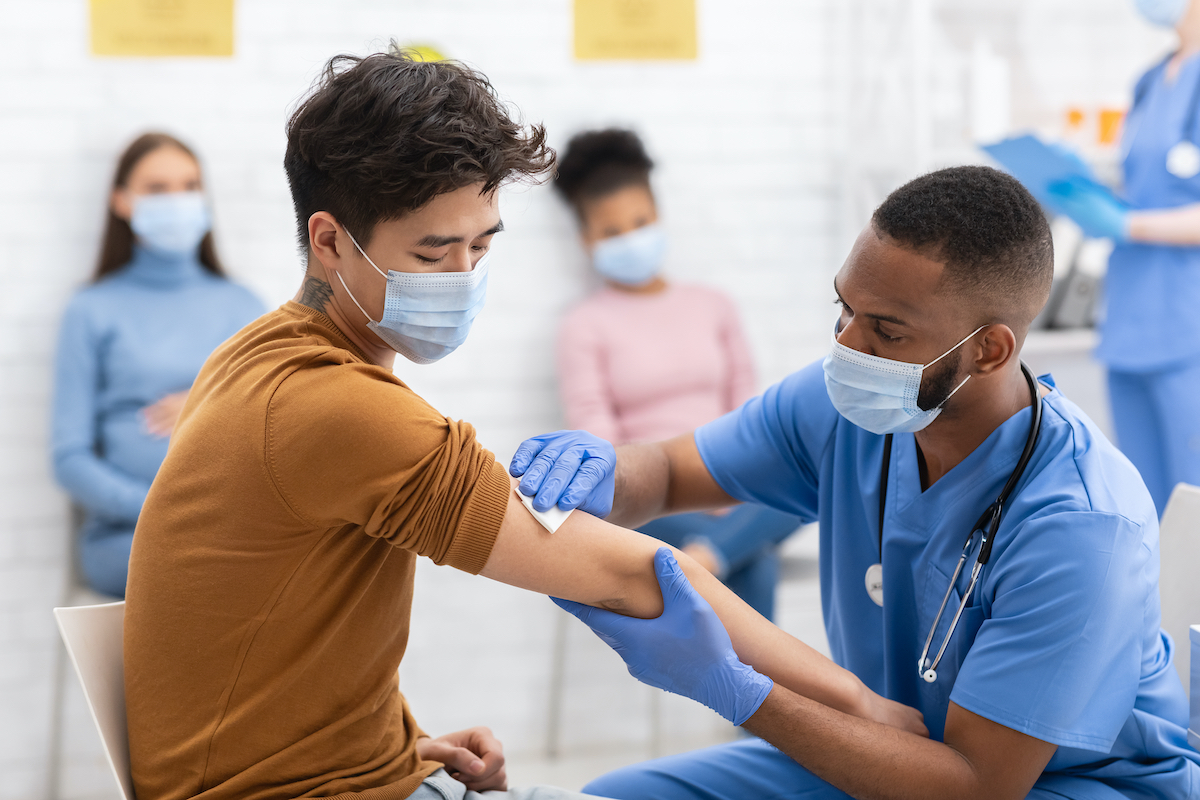 a black health professional kneels next to an asian man and presses a piece of gauze on his arm where he's just administered the covid-19 vaccine. both men are wearing masks and there's a line of people behind them