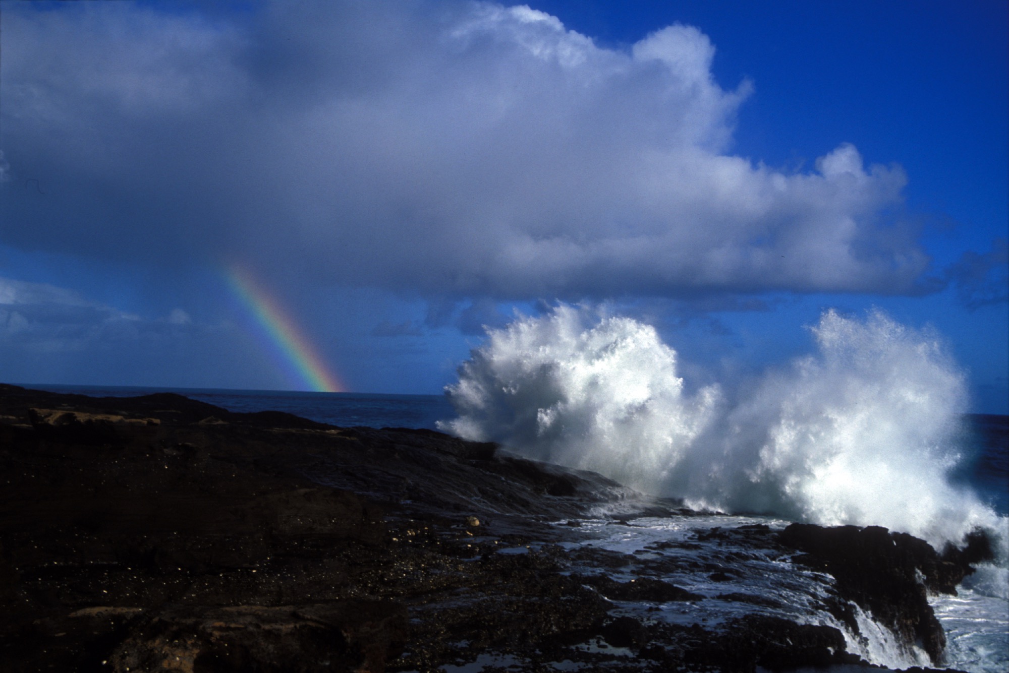 waves crash on a rocky shore. in the distance are patchy clouds and a pillar of a rainbow coming down from the cloud and disappearing into the ocean