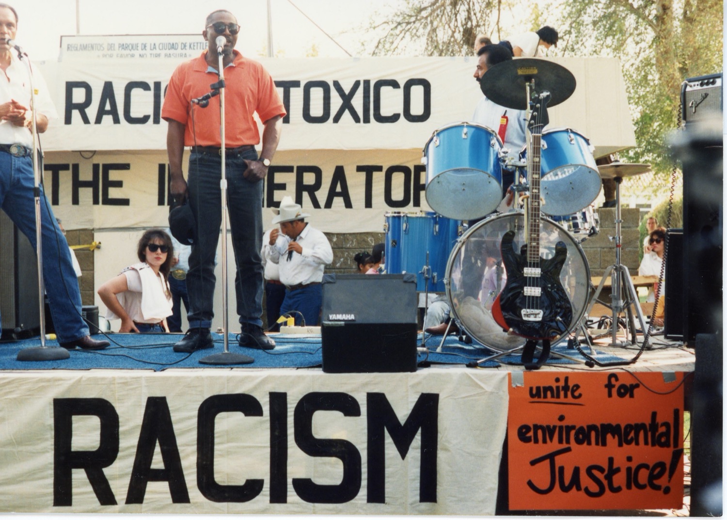 a black man in a red polo stands up on a stage and speaks in a mic at a protest rally. next to him are a band's instruments. signs read "toxico" and "racism" and "unite for environmental justice"