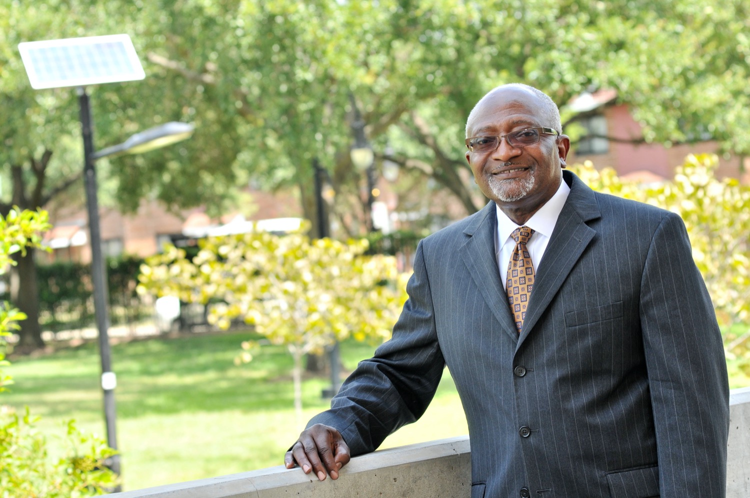 a profile headshot of a black man with glasses in a suit with trees in the background