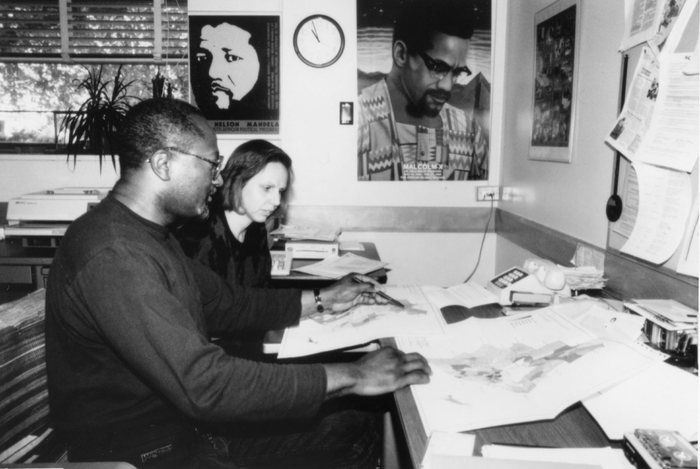 a black and white photo of a black professor teaching a graduate student. they are studying printout maps together