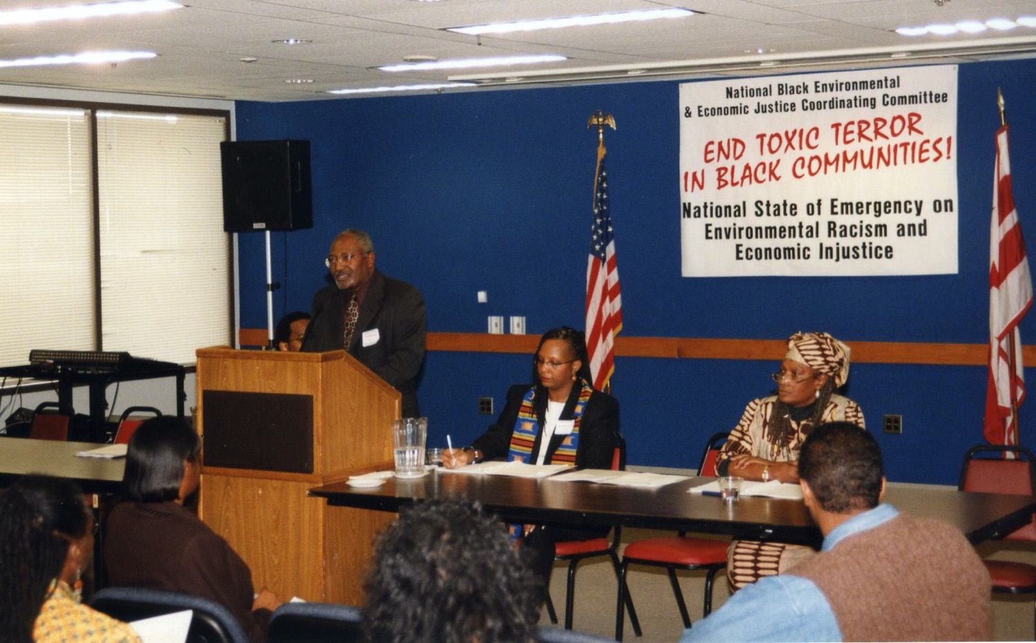 a black man speaks at a podium. next to him are two other black leaders at a panel table. behind them is a sign that can be partially read "end toxic... in black communities."