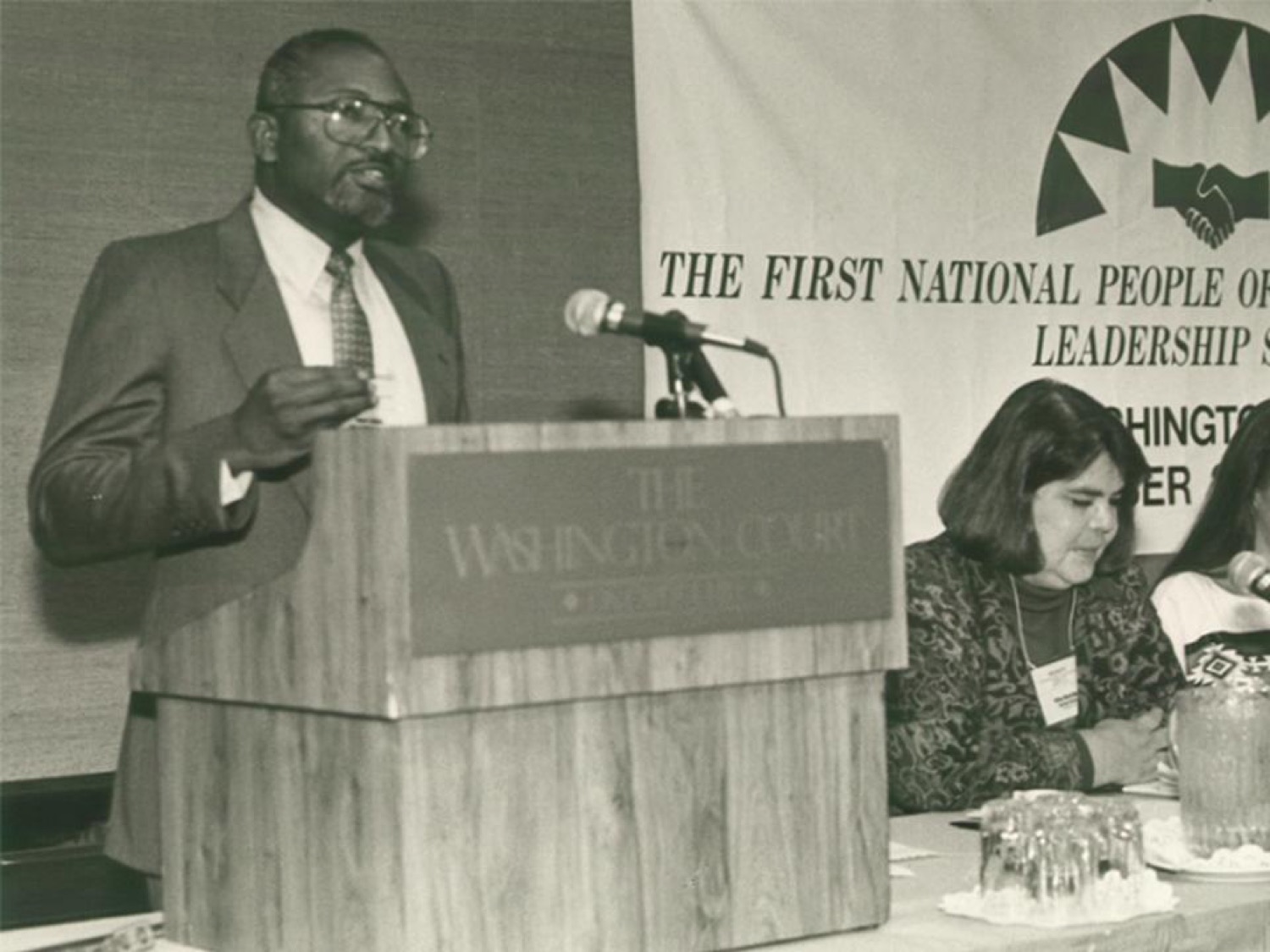 a black and white image of a black man in a suit speaking at a podium in front of a sign that said the irst National People of Color Environmental Leadership Summit