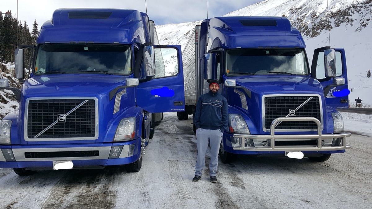 a man wearing a beanie, jacket, and sweats standing between two large blue delivery trucks in a snowy mountainous road