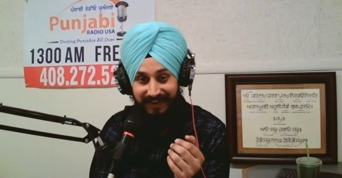 a man with a beard and wearing a blue turban speaks through a studio microphone and wears headphones