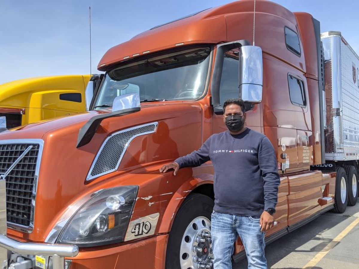 a man in jeans, a long sleeve shirt, and face masks stands in front of a large orange transportation truck.