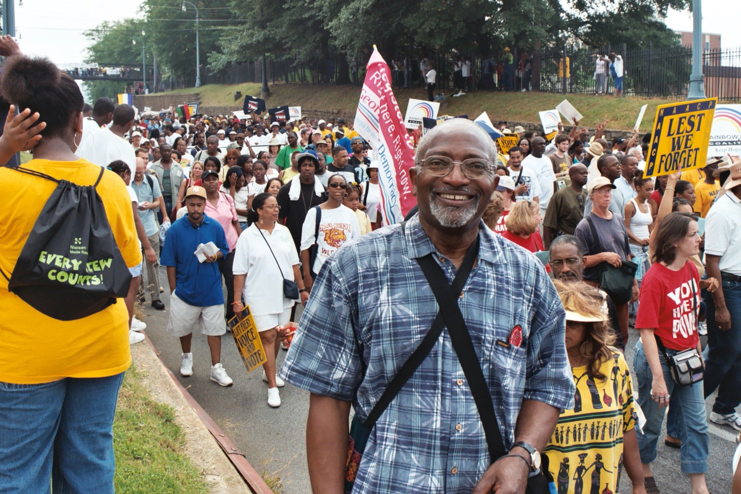 a black man in a blue plaid shirt smiles at the camera as he marches with a large crowd of marchers