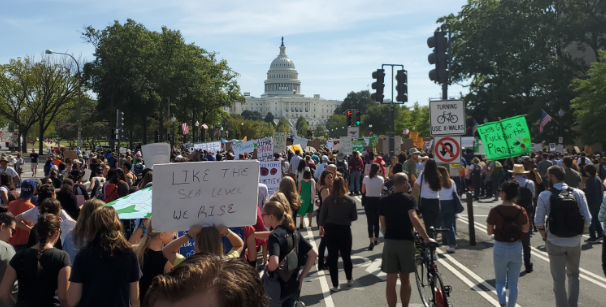 a large crowd of climate protesters carrying signs with slogans like "like the sea levels we rise" marching towards the capitol in dc