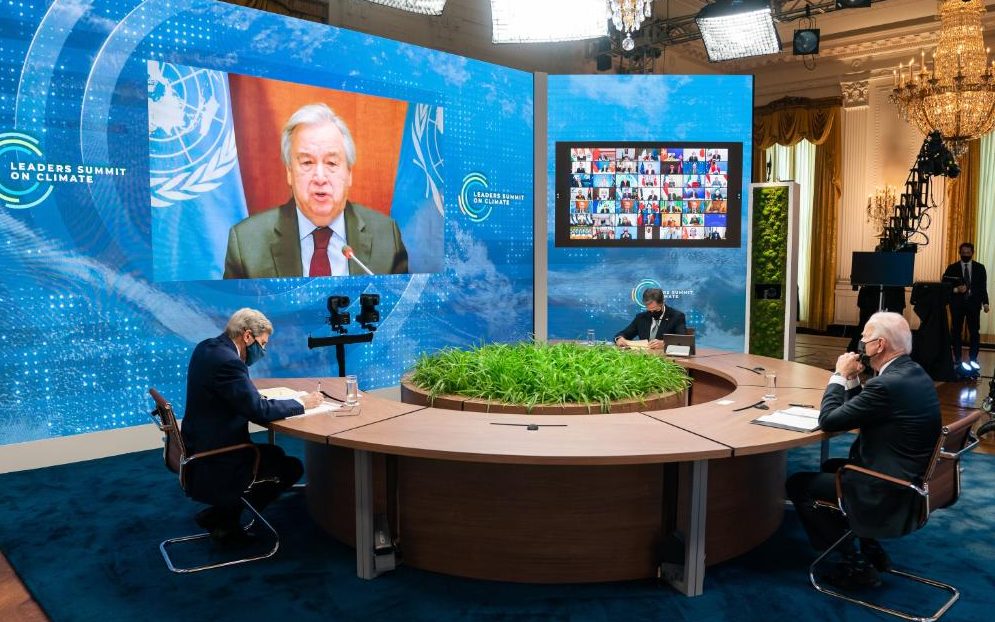 three men in face masks sit around a circular table and speak with people on a zoom call up on a large screen