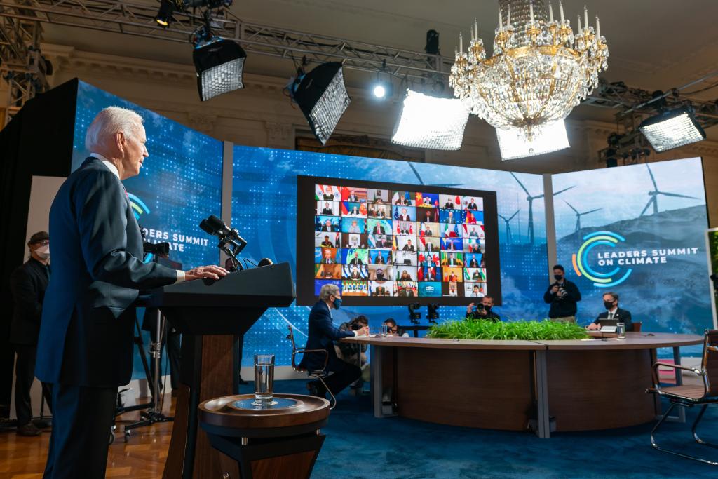 a man speaks at a podium behind to the side of him is a large screen with zoom callers and a board with a logo that reads leaders summit on climate