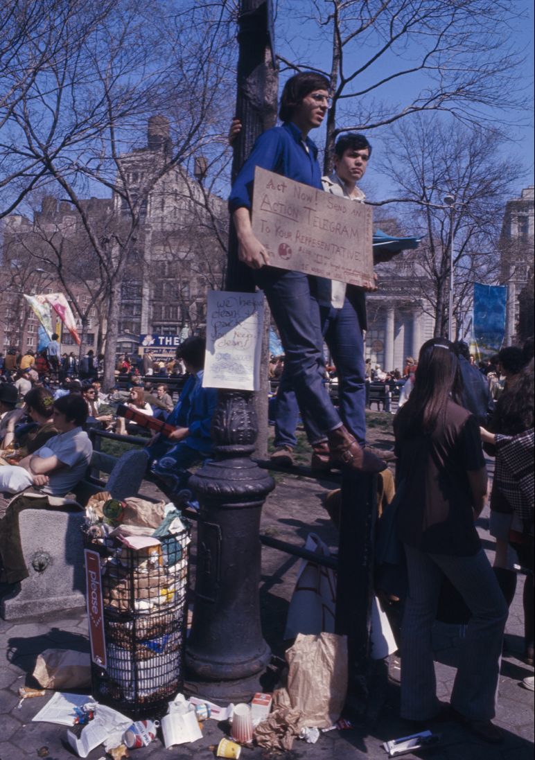 a crowd of people in new york, with the focus on a man in a blue shirt and glasses holding a cardboard sign that says "act now." he and another man are standing up against a tree. next to them is a trash can completely overfilled with trash 