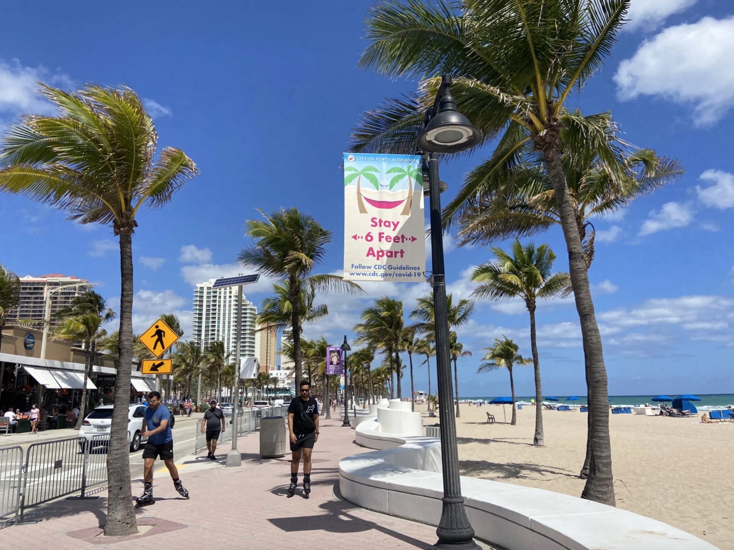 a daytime shot of the fort lauderdale boardwalk with palm trees and people rollerblading. a sign hangs from a lamppost saying 'stay 6 feet apart, follow cdc guidelines'