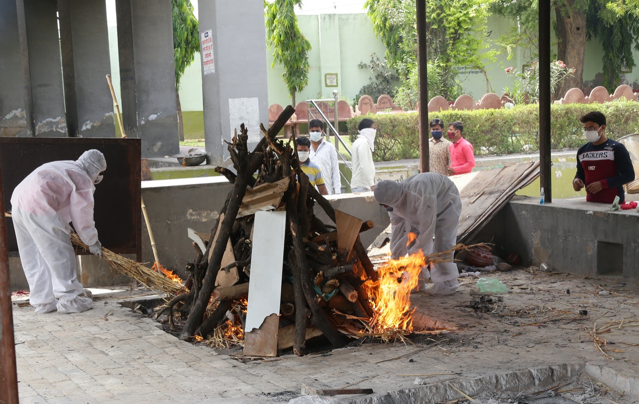 two people in hazmat suits attends to bonfire that is cremating someone who'd died from covid-19 complications in an abandoned building. people wearing masks observe from a distance