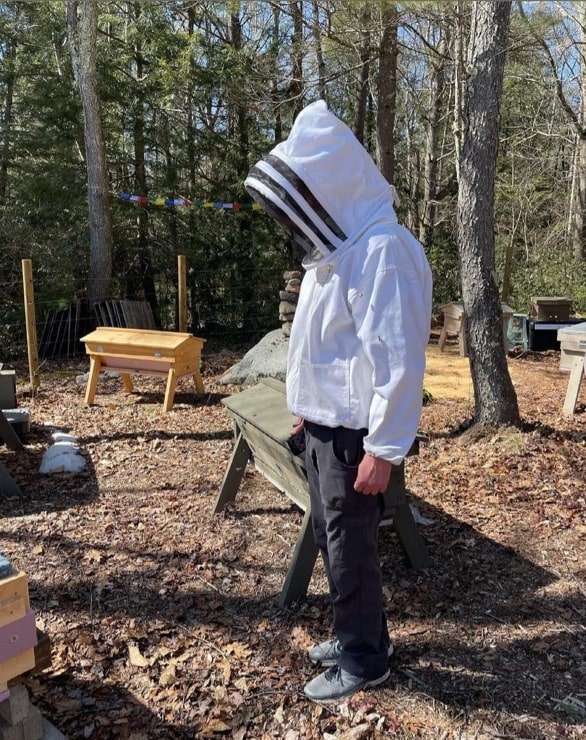 a figure stands in jeans and a white beekeeping top with a hood and veil, against a forest backdrop