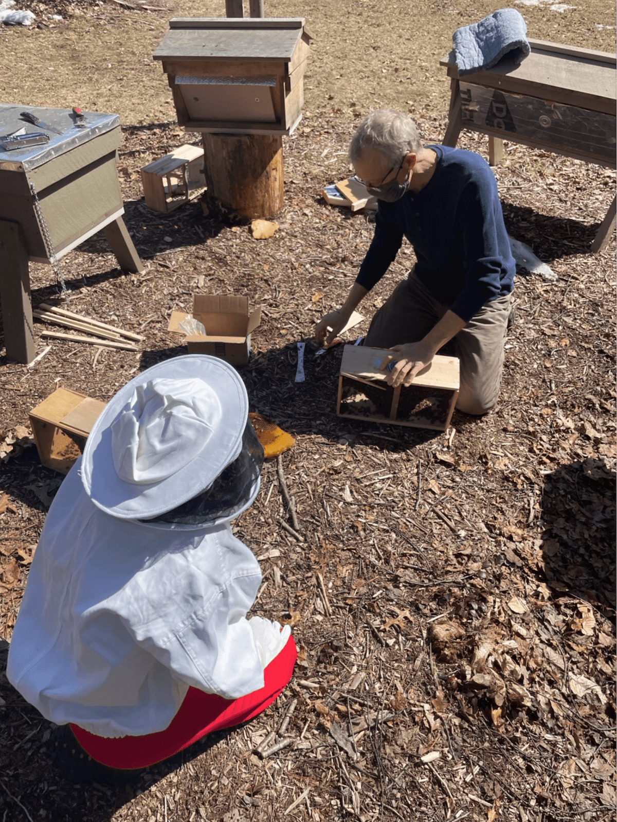 two people - one wearing a beekeeping suit and one in plain clothes - crouch over beekeeping materials in the dirt