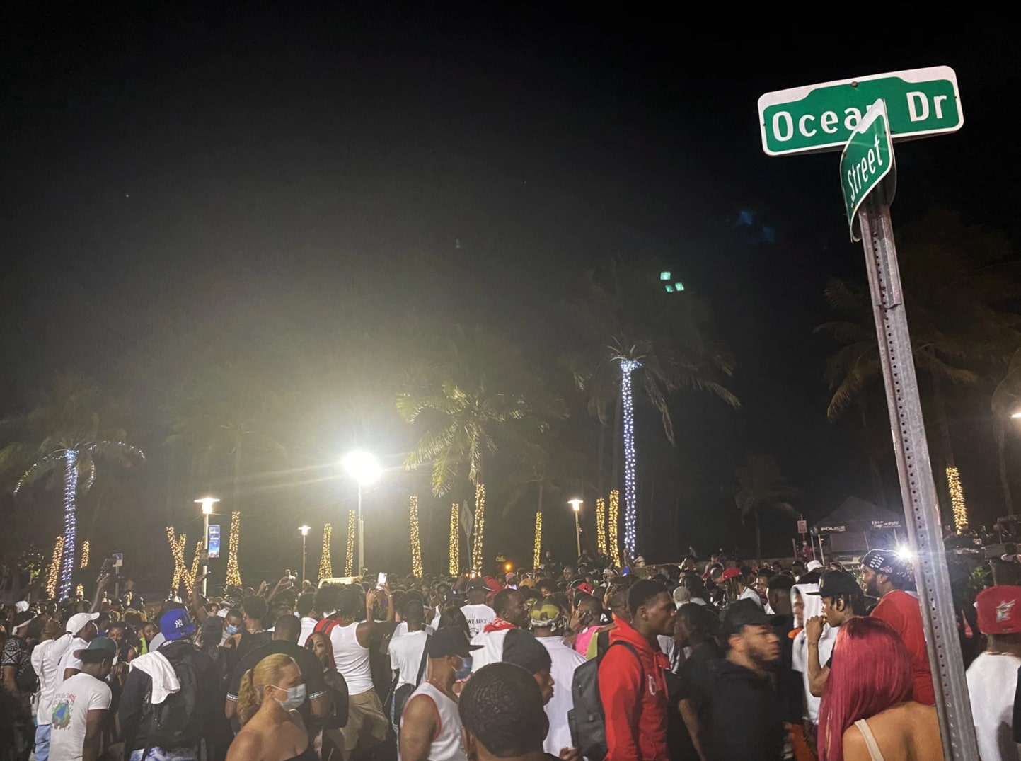 a large throng of people at night, tightly packed without masks. a street sign is in the foreground with palm trees decorated with string lights in the background