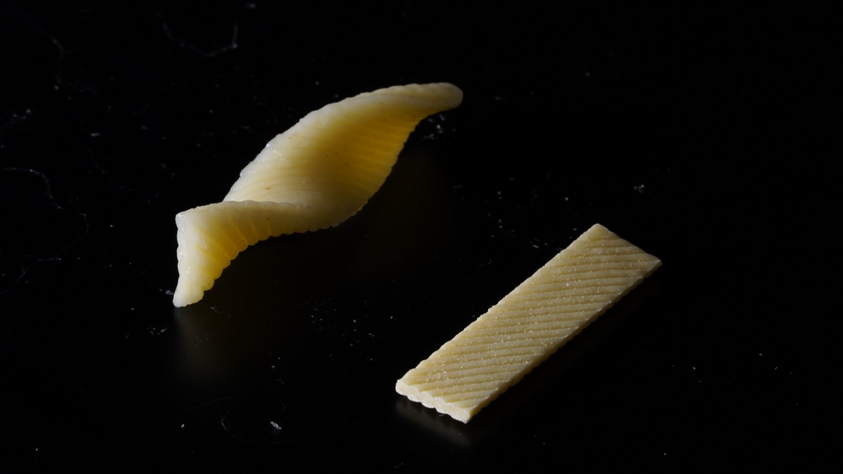 two pieces of pasta. in the right is a flat rectangular pasta. on the left is that pasta after being boiled in a twisted helix shape