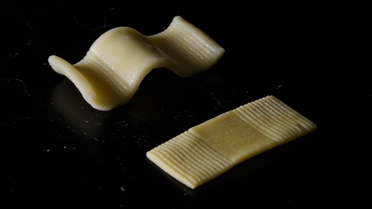 two pieces of pasta. on the right is a flat rectangular noodle. on the left is a that pasta after boiled showing a wavy shape