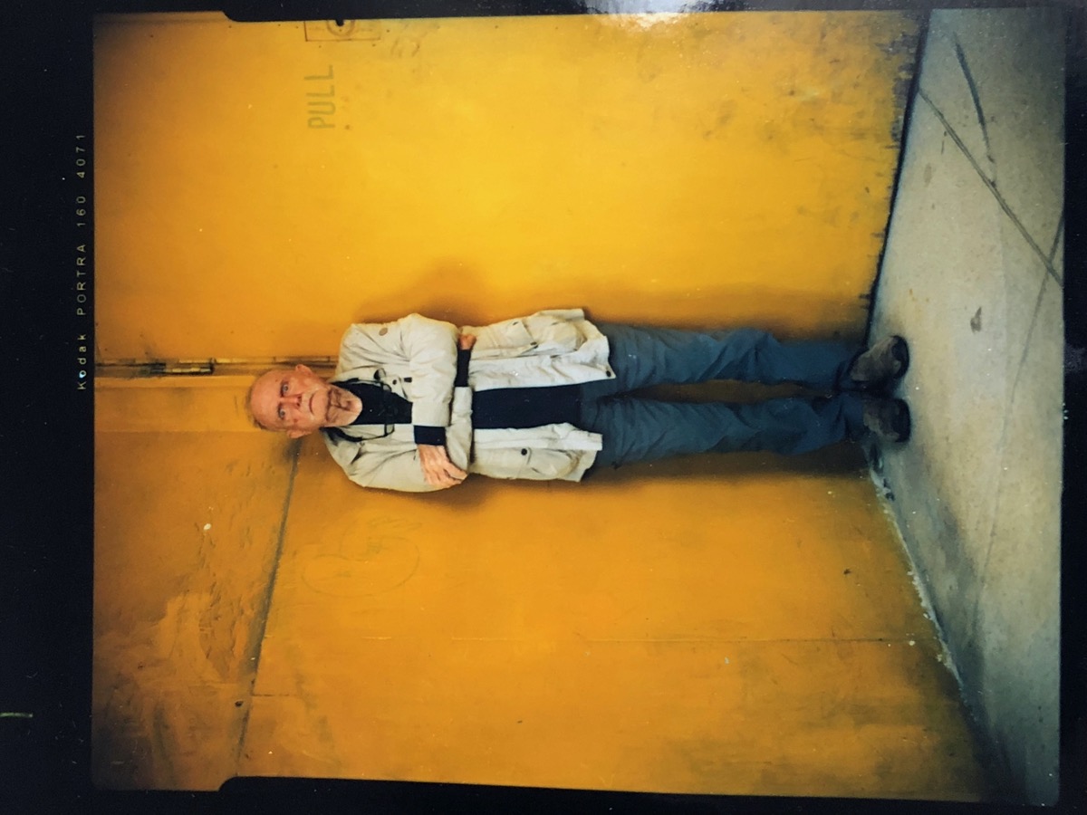 an older man in a denim shirt standing against a yellow background