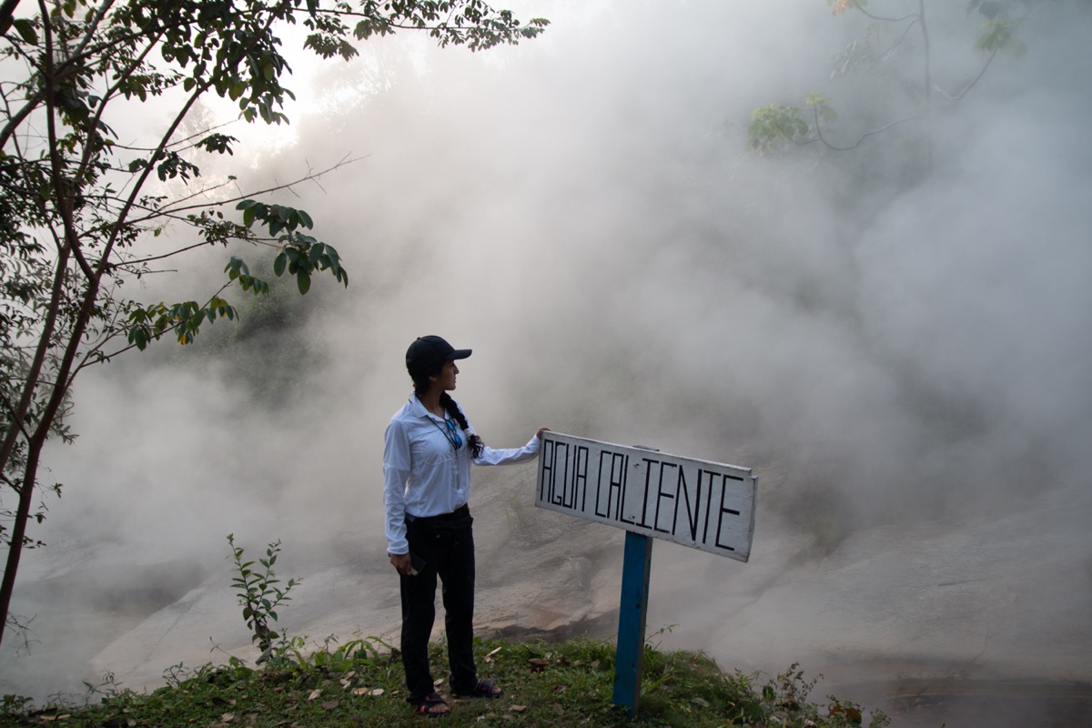 a woman scientist wearing gloves looks at a steaming river next to a sign in Spanish labeled "AGUA CALIENTE" (hot water)