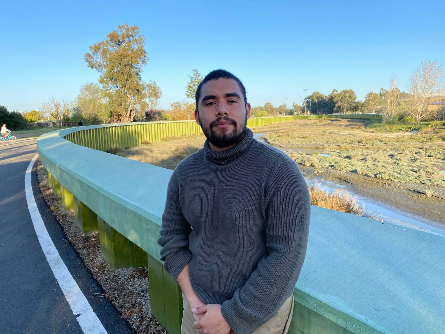 a man with a beard wearing a gray sweater leaning against a railing along the road