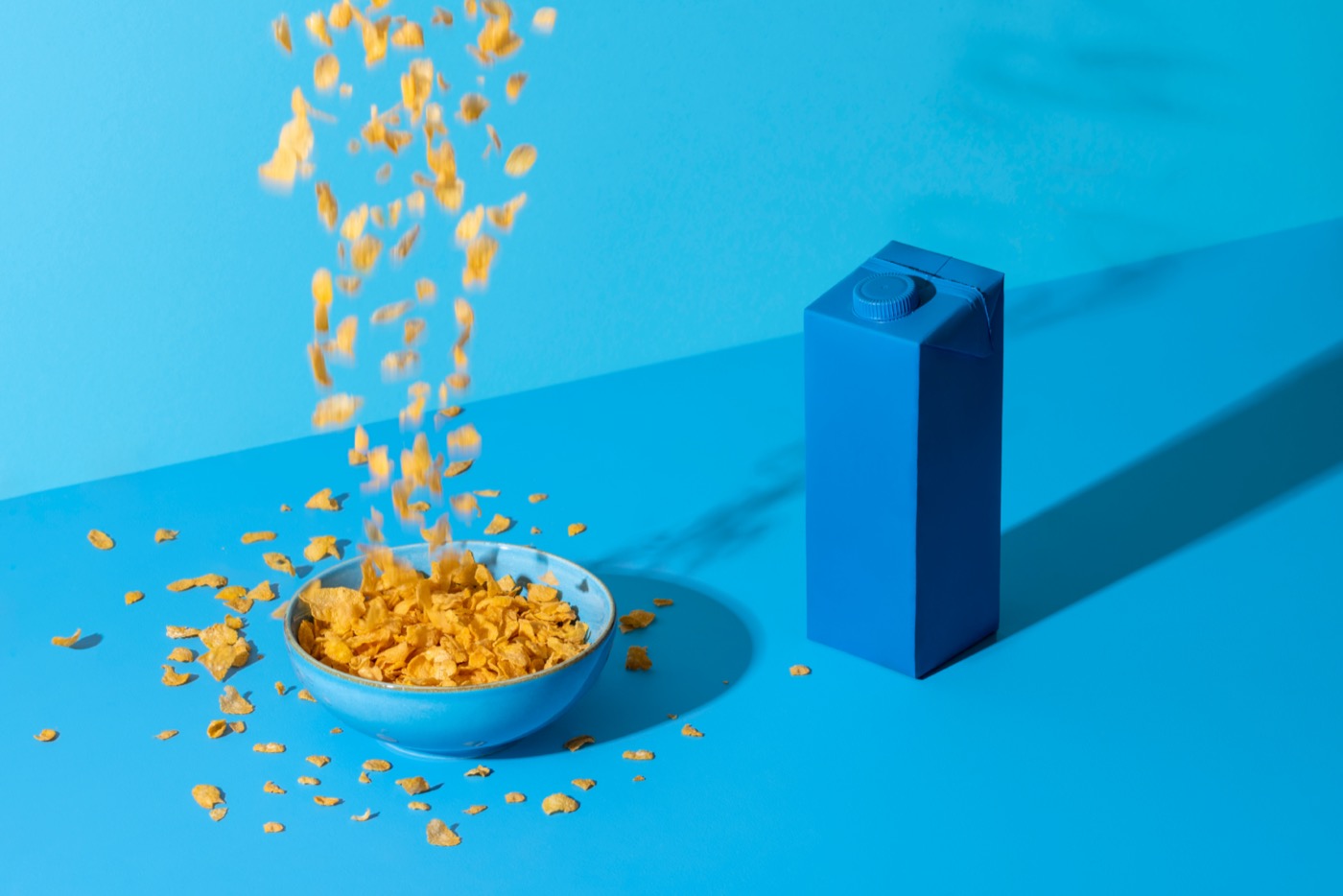 a waterfall of corn flakes falling into a blue bowl on a blue table with a blue carton of milk nearby