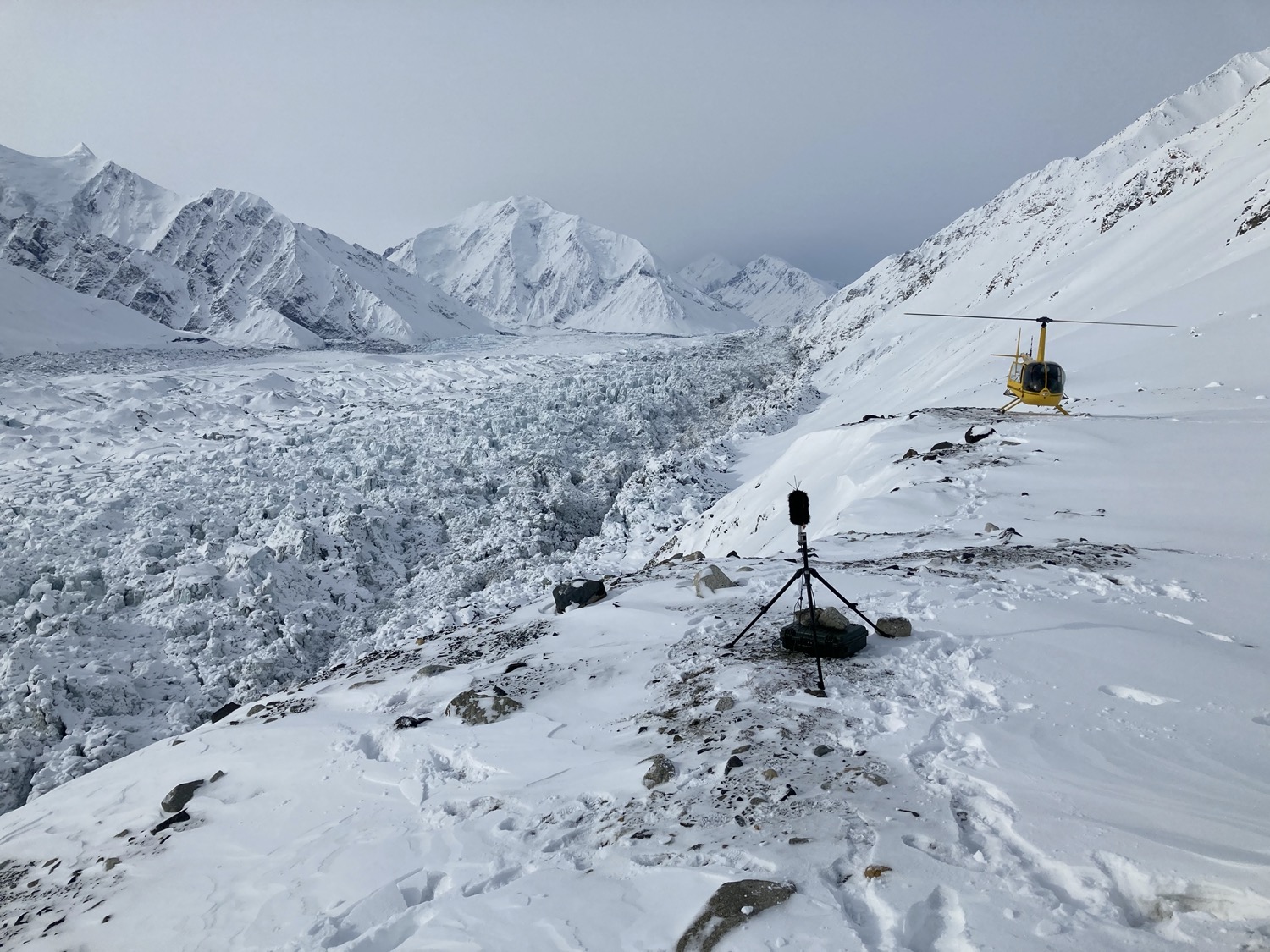 a snowy mountainous landscape, with a microphone on a tripod and a yellow helicopter in the background