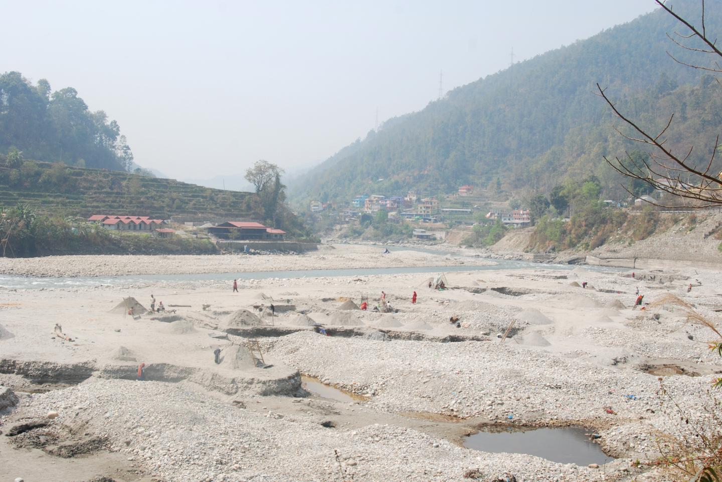 a sandy bank near a small stream. in the background are tree covered mountains and some small buildings