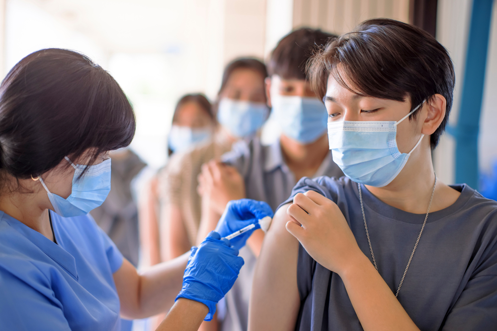 a teenaged boy with a face mask receives a vaccine shot in his arm by a medical provider in a mask and gloves. there are students lined up behind him