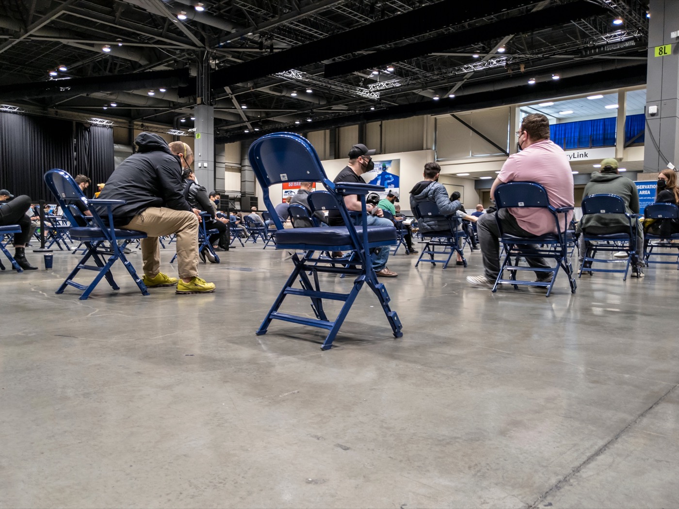 people sitting in rows of seats in large indoor stadium wearing masks. a chair in the foreground is empty