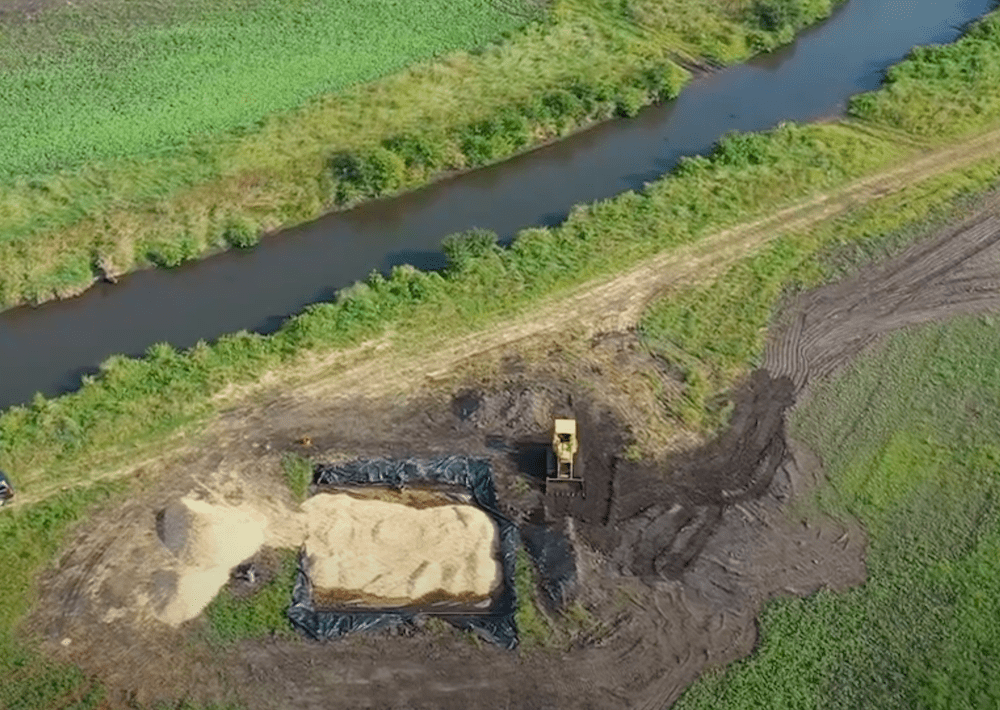 an aerial view of a large pile of woodchips and a trench of them with a tractor nearby. close to the trench is a small river