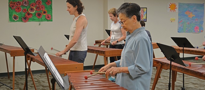 a group of people playing xylophones in a class, with the main focus on an older asian woman with short hair