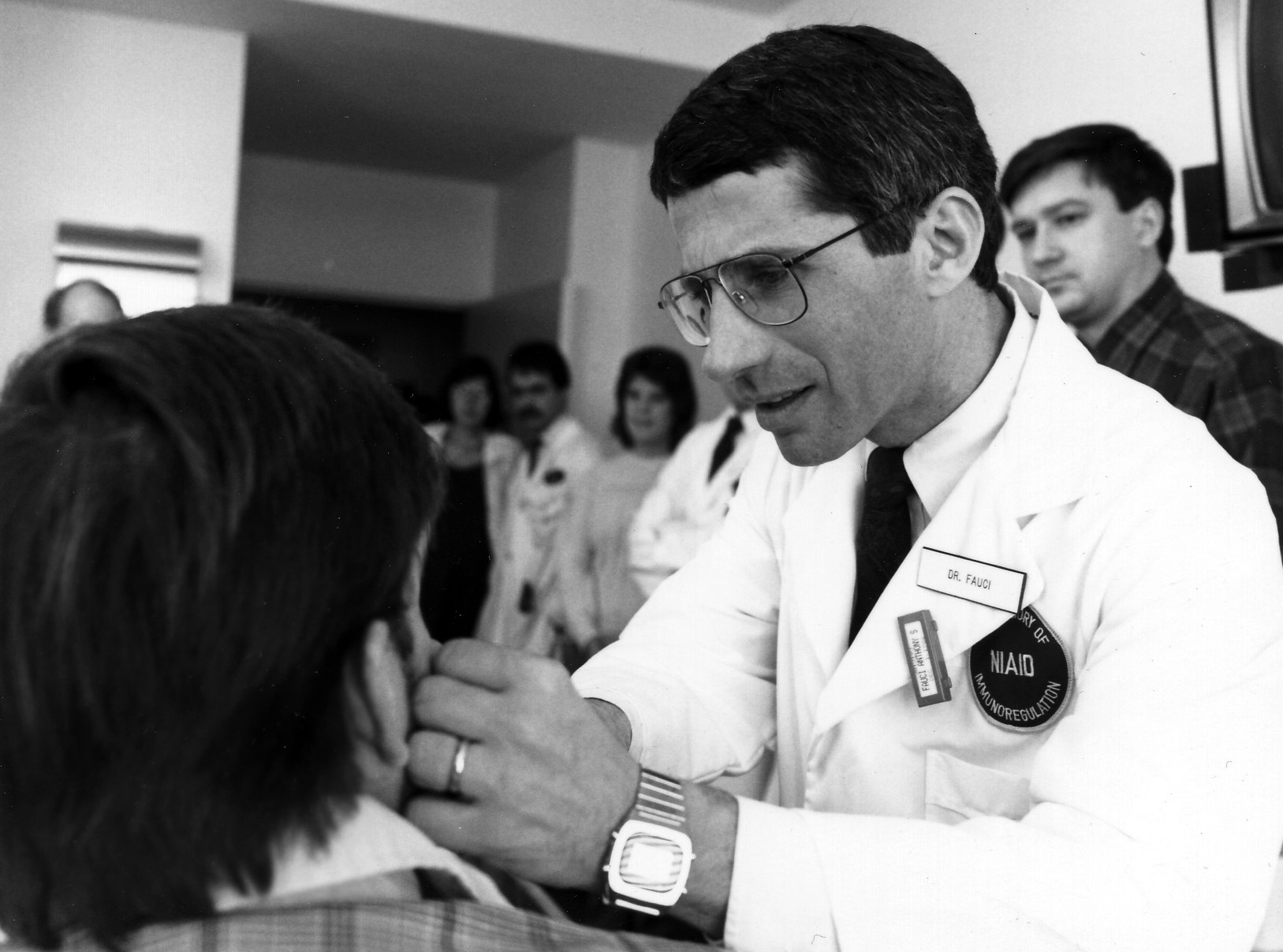 a black and white photo of a male doctor with glasses wearing a white coat with a patch that reads NIAID is examining a patient whose back is to the camera. other clinicians are in the office watching him treat the patient
