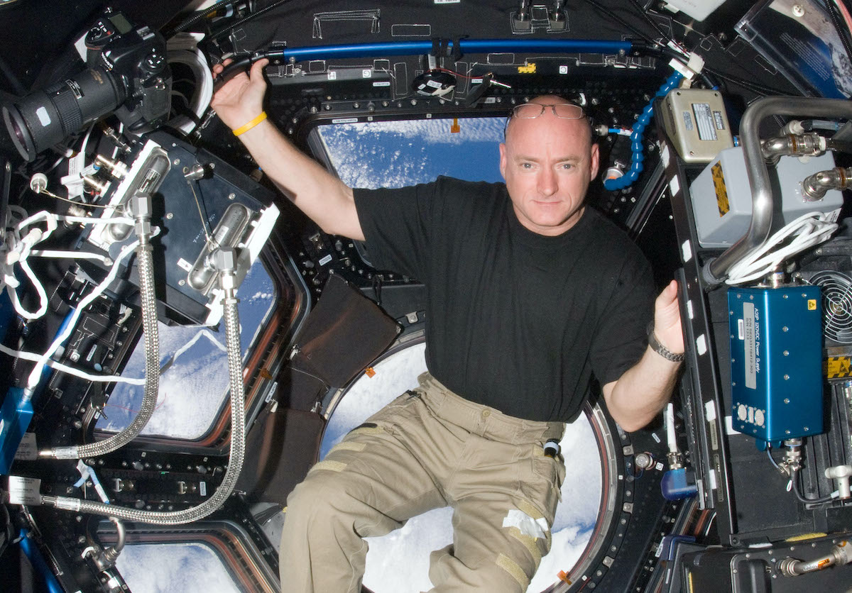 a white bald man in a black shirt and cargo pants floats in zero gravity inside a space craft that looks out over the earth