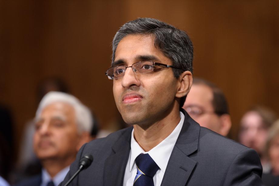 an indian man wearing glasses and a suit tie with a small microphone in front of him, with a small crowd of people behind him