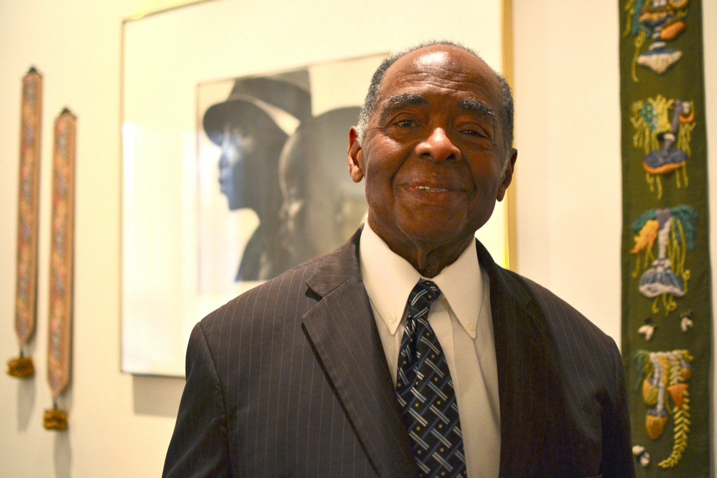 a black man in a suit with a large framed photograph behind him smiles at the camera