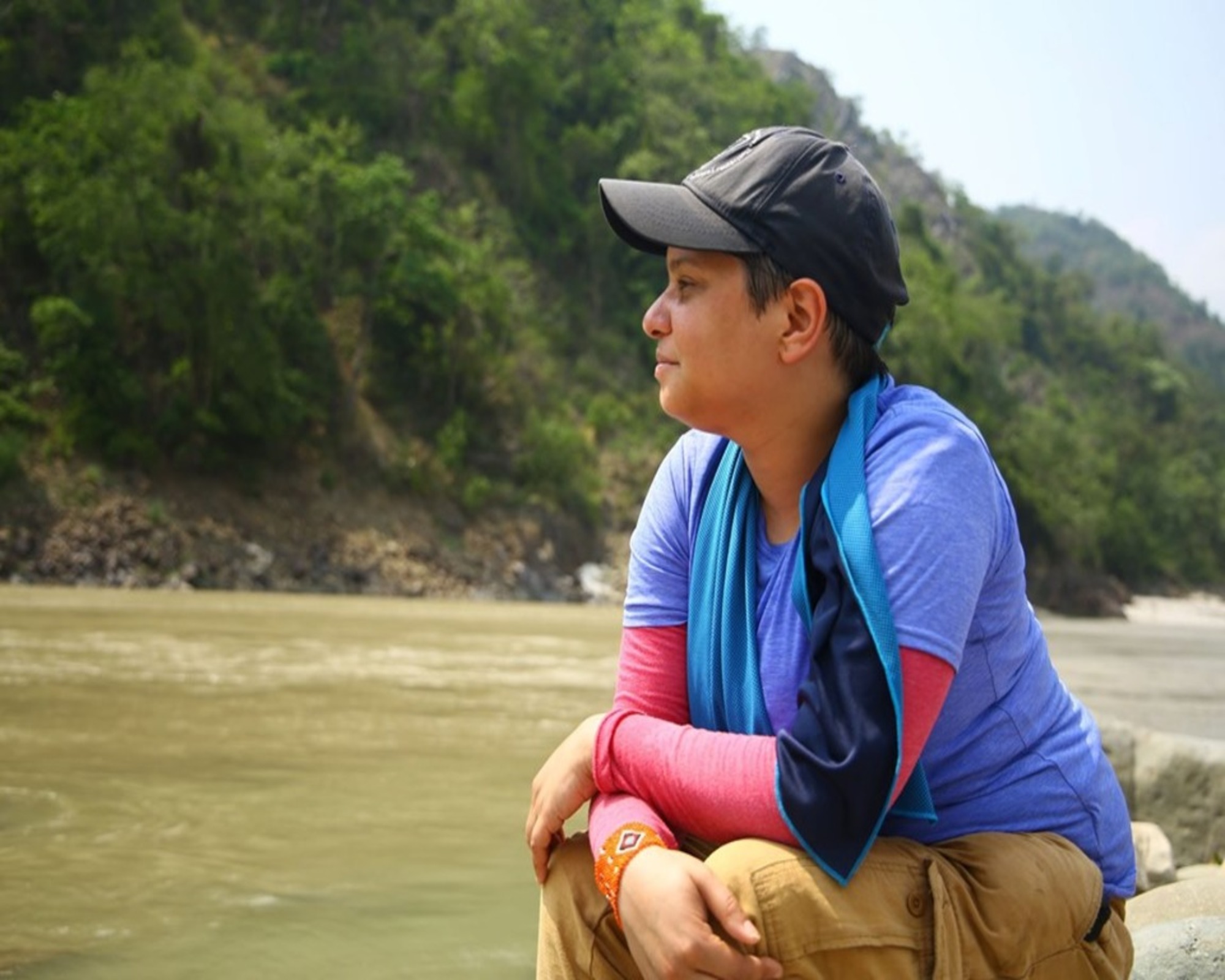 a nonbinary person wearing a baseball cap and a blue shirt looking out on a river with lush green behind them