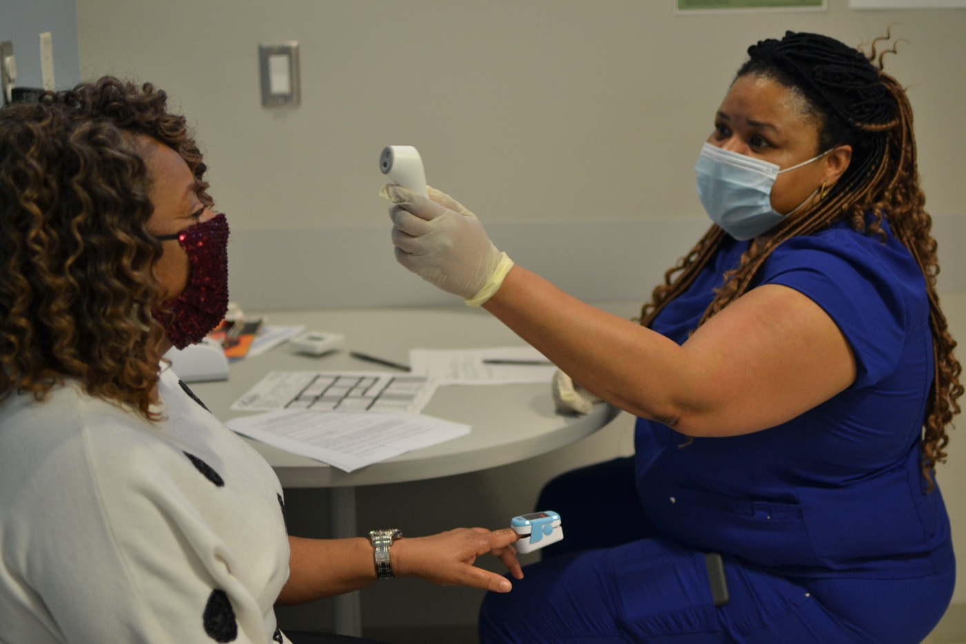 a black woman wearing scrubs and a mask, takes the temperature of another black woman, also wearing a mask