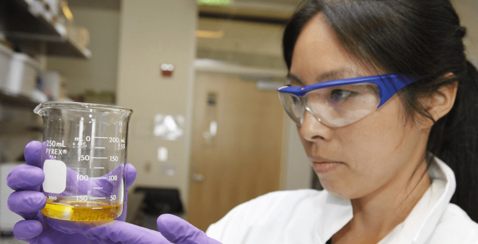 Dr. Selina Wang is wearing safety goggles and a white lab coat and purple latex gloves, and is holding up and looking into a beaker with dark liquid in it