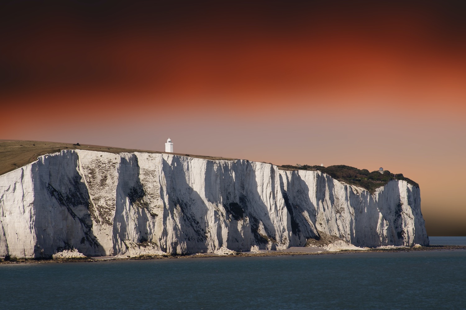 sheer cliffs the color of white chalk over the ocean. atop the cliffs is a lighthouse. the whole landscape is under a red, orange sunset