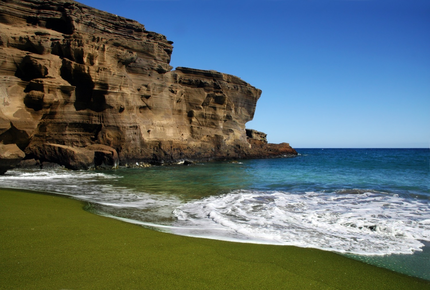 a beach with green sand that looks the shade of moss. in the horizon are weathered cliffs