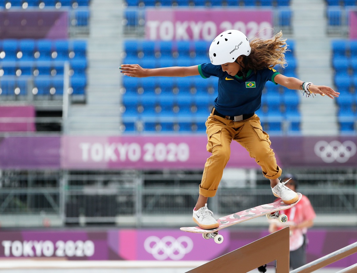 a young brazilian girl in a white helmet, navy polo shirt, and khaki cargos rides a pink and black skateboard over a railing. in the background is an empty auditorium with signs for the tokyo 2020 olympics