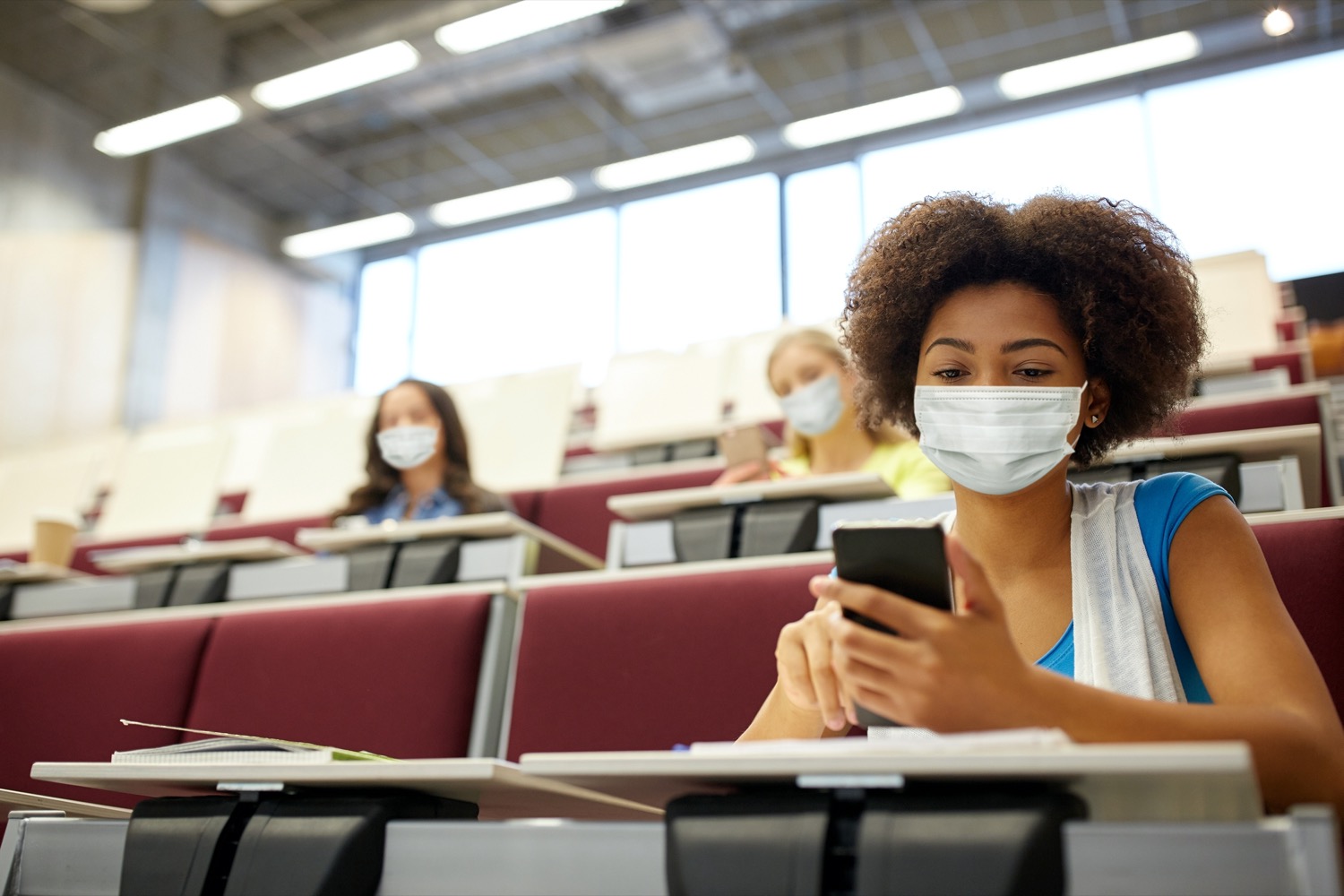 three teen students in a classroom all wearing masks. in the foreground a black girl looks at her phone